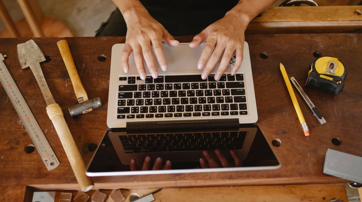 Mains sur le clavier d'un ordinateur portable posé sur un établi en bois, entouré d'outils d'artisan, pour illustrer l'accessibilité web artisan