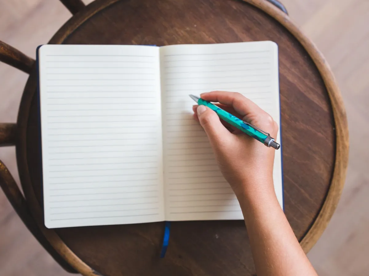 Main qui note une liste de démarches dans un carnet ouvert sur un tabouret en bois