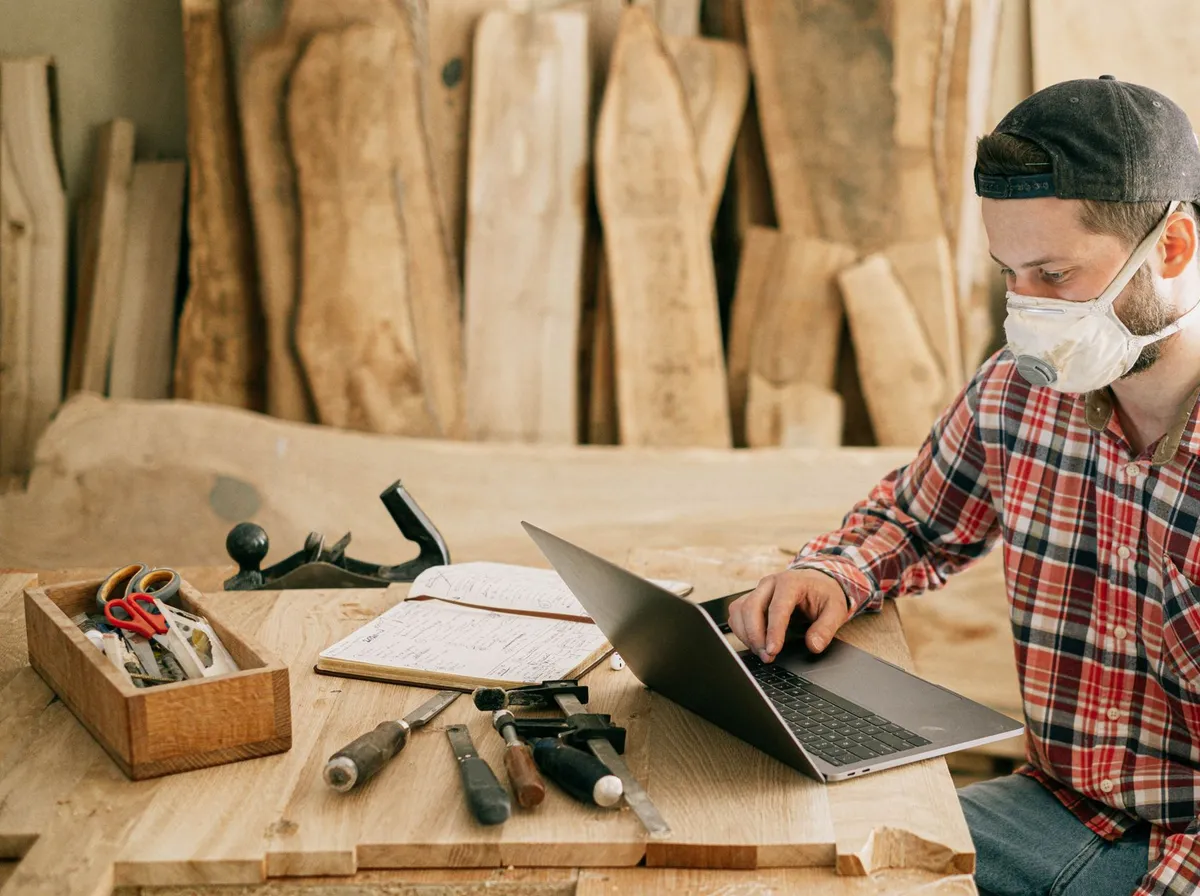 Artisan travaillant sur un ordinateur portable posé sur son établi, entouré de ses outils et d'un carnet de croquis, atelier bois du Finistère