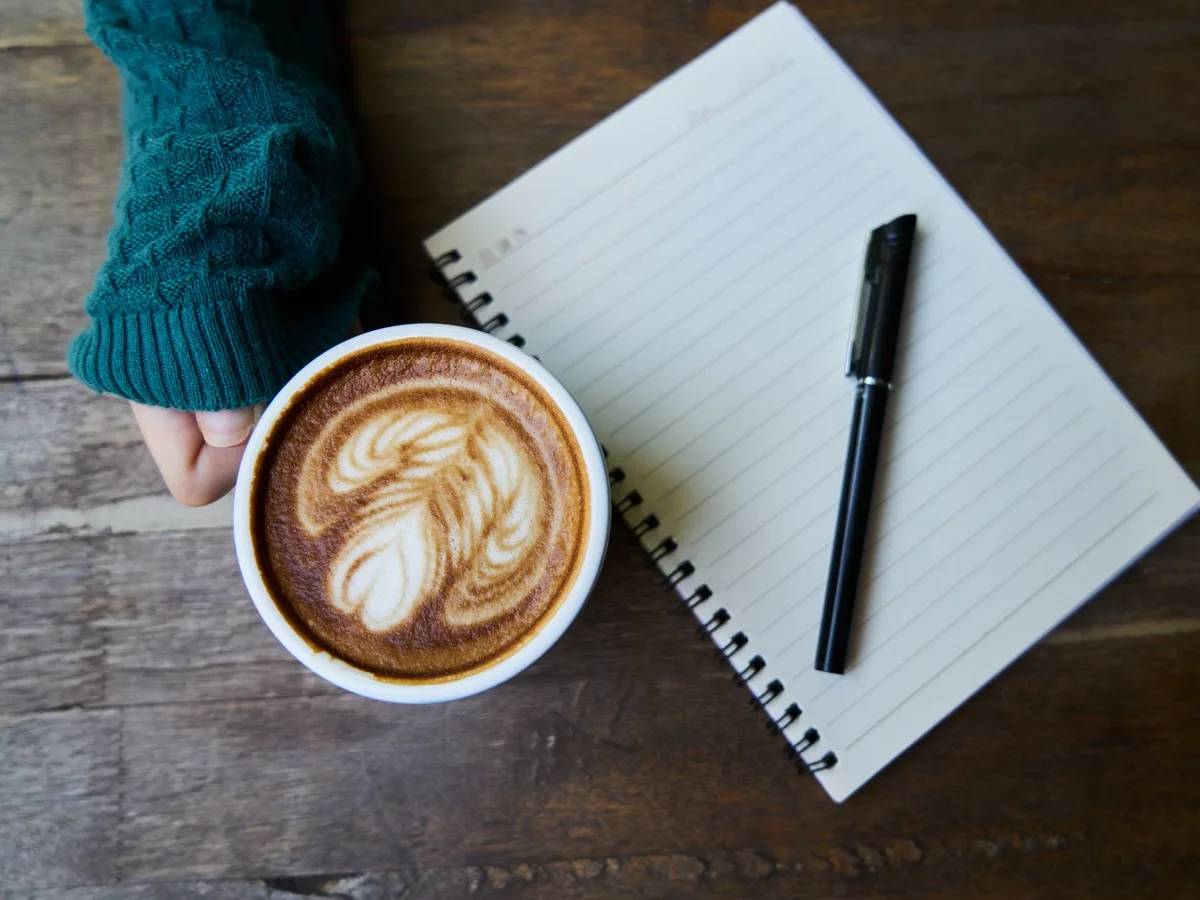 Tasse de café et carnet vierge ouvert avec un stylo, posés sur un bureau en bois sombre, avant de rédiger une réponse à un avis négatif