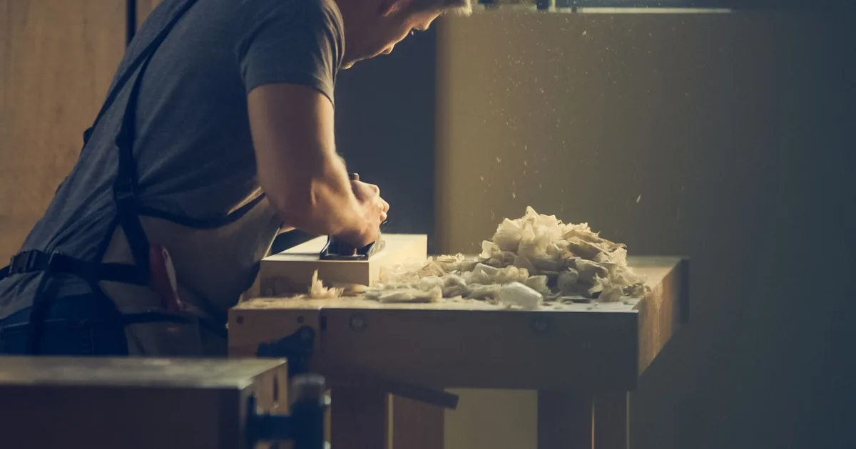 Artisan menuisier de profil rabotant une pièce de bois dans son atelier, copeaux en lumière chaude