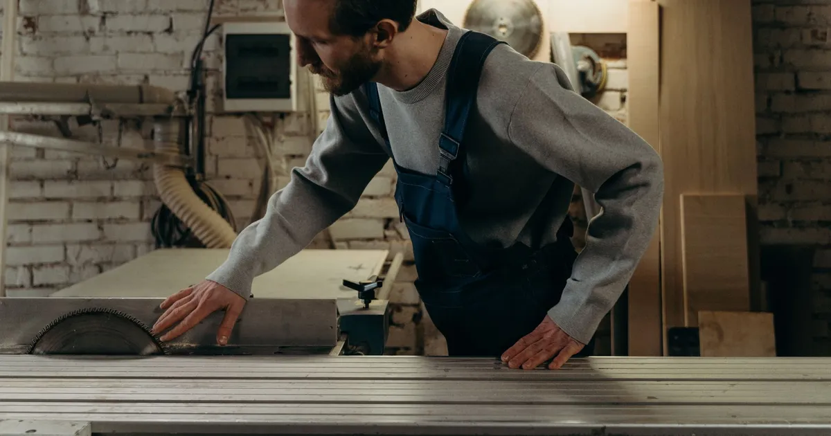 Menuisier de profil dans son atelier, la main posée près de la scie à table, murs de brique blanche