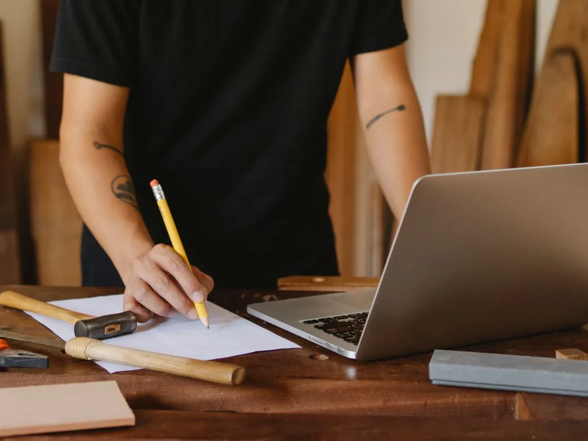 Mains annotant un croquis à côté d'un ordinateur portable sur un établi en bois, préparation d'un site internet d'artisan